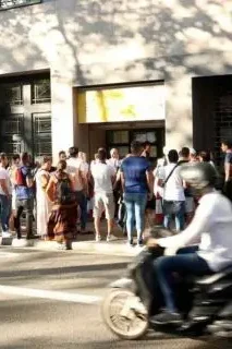 Grupo de personas haciendo fila frente a un edificio, con una moto pasando por la calle.