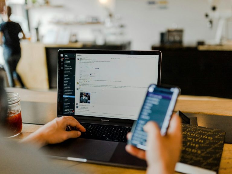 Persona trabajando en una laptop mientras sostiene un teléfono móvil en un café.