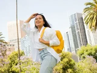 Mujer sonriente con mochila amarilla frente a edificios y palmeras.