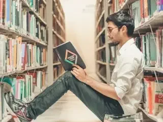 Joven leyendo un libro en una biblioteca, sentado entre estantes de libros.