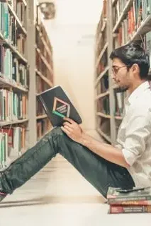 Chico sentado en una biblioteca, leyendo un libro entre estanterías de libros.